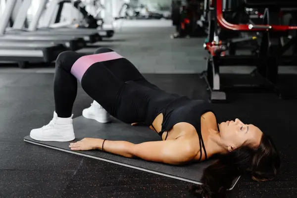 Young woman in black sportswear training with elastic resistance band above knees in gym. Lady doing glute bridge exercise for buttocks, legs and core.